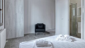 A modern hotel room with a large wardrobe, a black chair, a neatly made bed with towels, and a glass shower area. - Suite Hendrix | BluesHouse Carracci (Bologna)