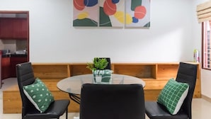 Un comedor con una mesa redonda de vidrio, dos sillas negras con almohadones verdes y una obra de arte abstracta en la pared.