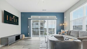 Ein modernes Wohnzimmer mit einem grauen Ecksofa, einem langen, grauen Konsolentisch, einem flachbildfernseher an der Wand und einem großen Fenster mit Jalousien.