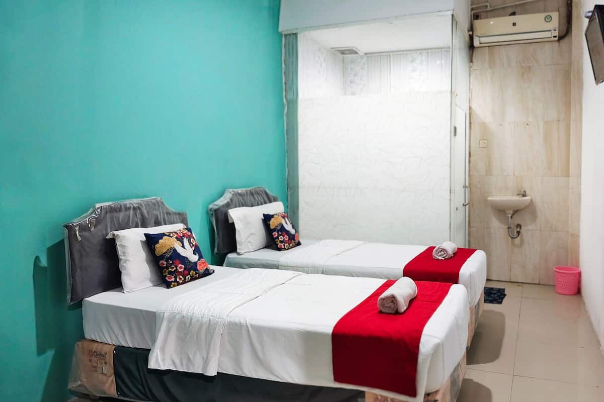 Ein Zimmer mit zwei Betten, jeweils mit einer roten Decke und einem Kissen mit einem Blumenmuster. In der Ecke befindet sich ein weißes Waschbecken und ein kleiner Mülleimer.