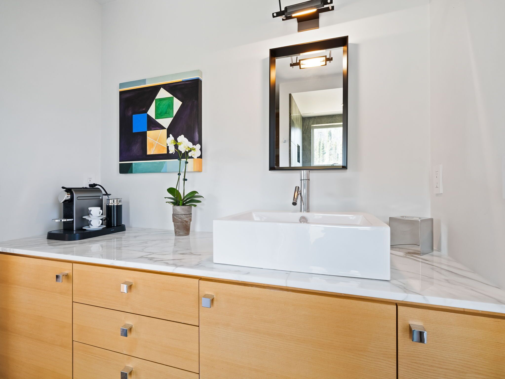 A modern bathroom with a white sink, a coffee machine, a potted orchid, and a geometric painting on the wall.