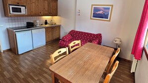 Une cuisine compacte avec des meubles en bois, un four à micro-ondes et une machine à café. On y trouve une table à manger avec des chaises, ainsi qu’un canapé rouge à motifs floraux. Un tableau encadré représentant une montagne est accroché au mur.