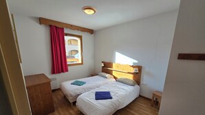Une chambre avec deux lits, un plancher en bois, une fenêtre avec des rideaux rouges, une tête de lit en bois et une petite table de chevet avec une lampe.