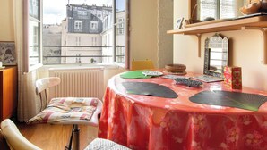 Ein Zimmer mit Blick auf Gebäude durch ein Fenster, ein Tisch mit rotem Blumenmuster bezogen, ein Stuhl und ein Spiegel an der Wand.