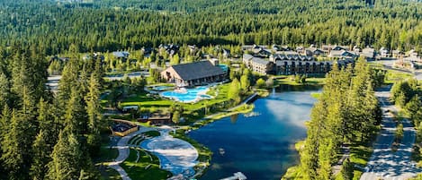 An aerial view of a resort with a large swimming pool, a dock, and a forested backdrop.