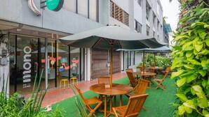 An outdoor seating area with wooden tables and chairs, a green awning, and a glass door entrance to a building with a 'mono living' sign. - Mono Coliving Kota Tua, Jakarta (Jakarta)
