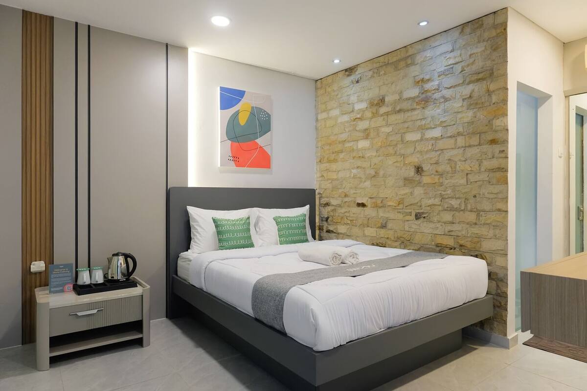 A modern bedroom with a stone accent wall, a bed with white bedding and green pillows, a bedside table with a coffee maker, and a framed abstract art piece above the bed.