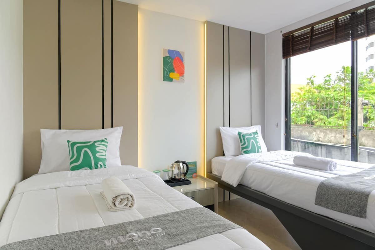 A hotel room with two beds, a bedside table with a kettle, a small artwork on the wall, and a window with a view of greenery.