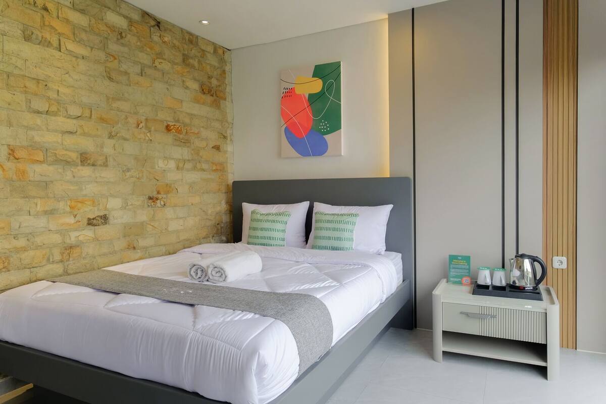 A modern hotel room with a brick wall, a large bed with white bedding, and a small bedside table with a kettle and cups.