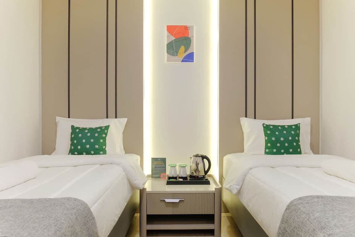 A hotel room with two beds, each with white bedding and green pillows with a circular pattern. There is a nightstand with a coffee maker, a kettle, and a sign. The room has a neutral color scheme with beige walls and a modern design.