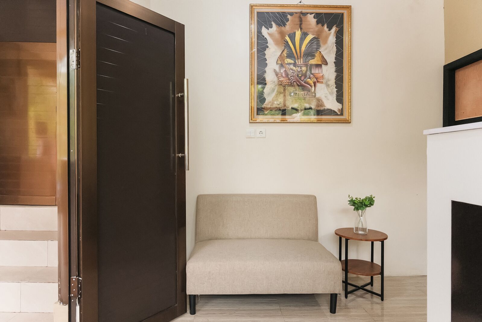 A room with a closed dark wooden door, a beige armchair, a small round table with a plant, and a framed artwork on the wall.