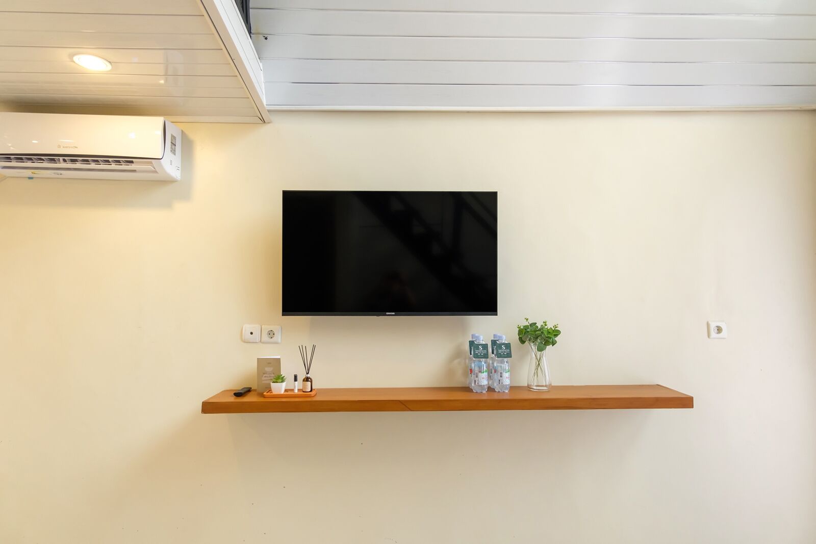 A modern living room with a mounted flat-screen TV, a wooden shelf with decorative items, and a wall-mounted air conditioner.