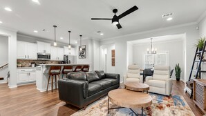 A spacious living area with a black leather sofa, a round coffee table, and a white armchair. There is a kitchen in the background with white cabinets and a microwave.