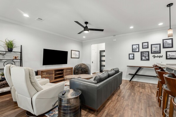 A modern living room with a grey sectional sofa, a white recliner, a flat-screen TV, and a ceiling fan.