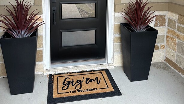 A black front door with three glass panels, flanked by two potted plants, and a doormat with the text "Gig' em THE WELLBORN'S".