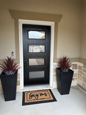 A black front door with three glass panels, flanked by two potted plants, and a doormat with the text "Gig' em THE WELLBORN'S".