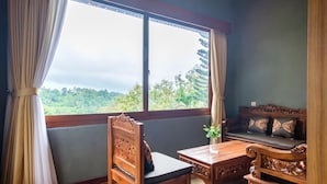 A room with a wooden table and chair, a cushioned bench, and a large window offering a view of greenery. - Calma Coliving at Jatiluwih, Bali (Penebel)