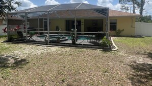 A covered outdoor swimming pool with a patio area, surrounded by a grassy yard and a house in the background. - Welcoming 3-bedroom house with pool and private yard (North Port)