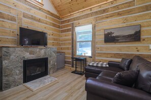 Ein Holzblockhaus-Interieur mit Kamin aus Stein, einem Ledersofa, einem Fernseher, der über dem Kamin angebracht ist, und einem Fenster mit Blick ins Freie.
