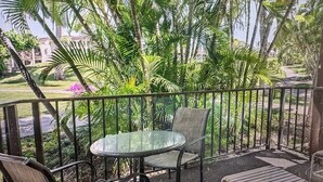 A covered outdoor area with a glass table and chairs, surrounded by lush greenery and a view of a building in the background.