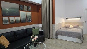 Una habitación moderna con un sofá de cuero negro, una mesita redonda con una planta en maceta, una cama con cubrecama gris y una pared con varios cuadros.
