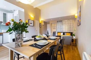 Un comedor moderno con una mesa de madera dispuesta para cuatro, un jarrón de flores y una cocina al fondo.