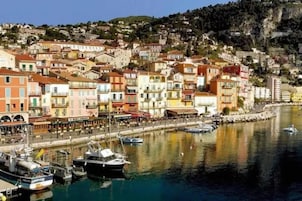 Un pueblo costero con edificios coloridos, un puerto deportivo con barcos y una colina al fondo.