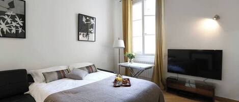 Une chambre à coucher avec un lit, une télévision, une petite table avec une lampe et des œuvres d’art accrochées au mur.