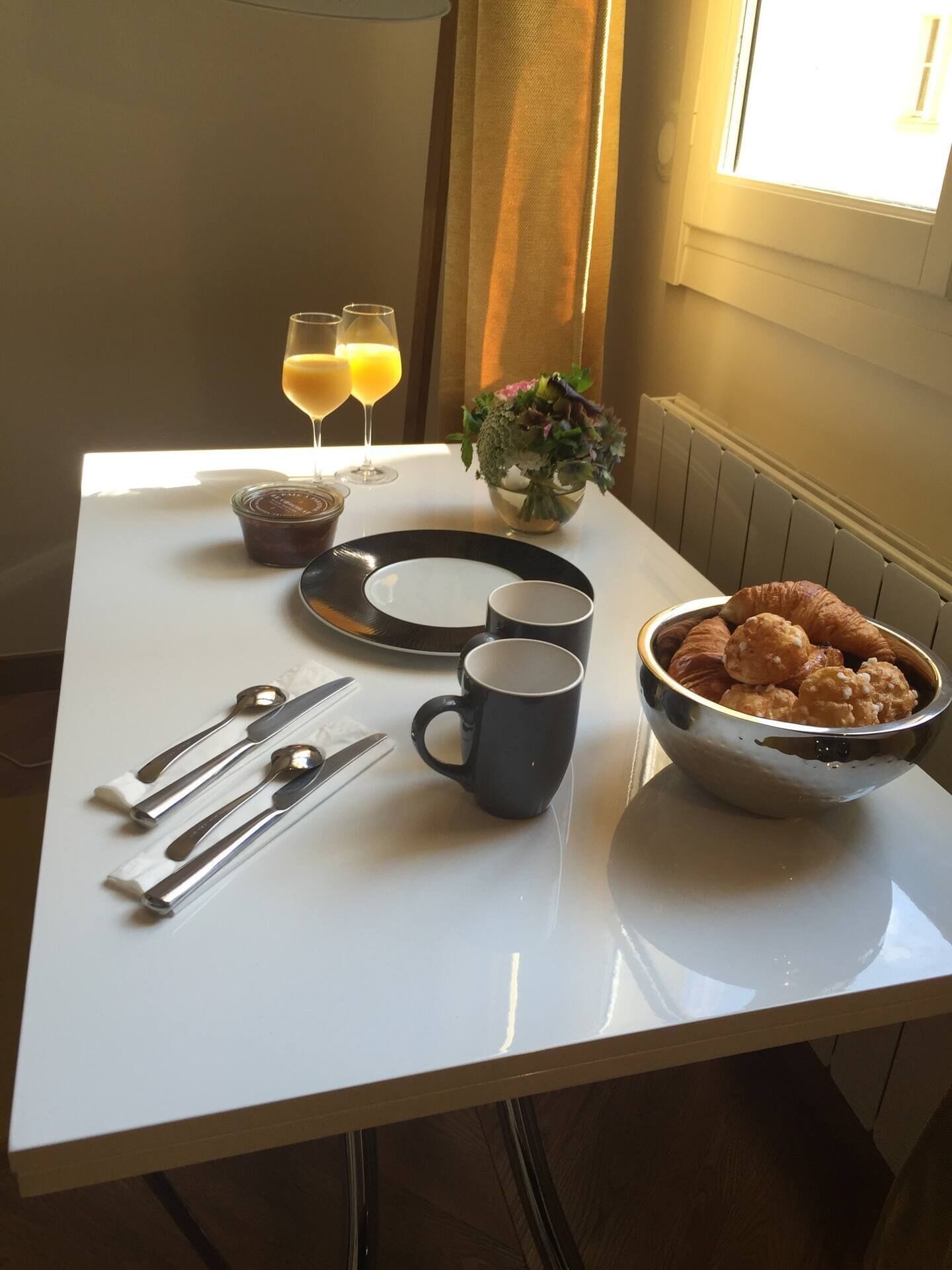 A breakfast table with a bowl of pastries, two glasses of orange juice, a vase with flowers, and cutlery.