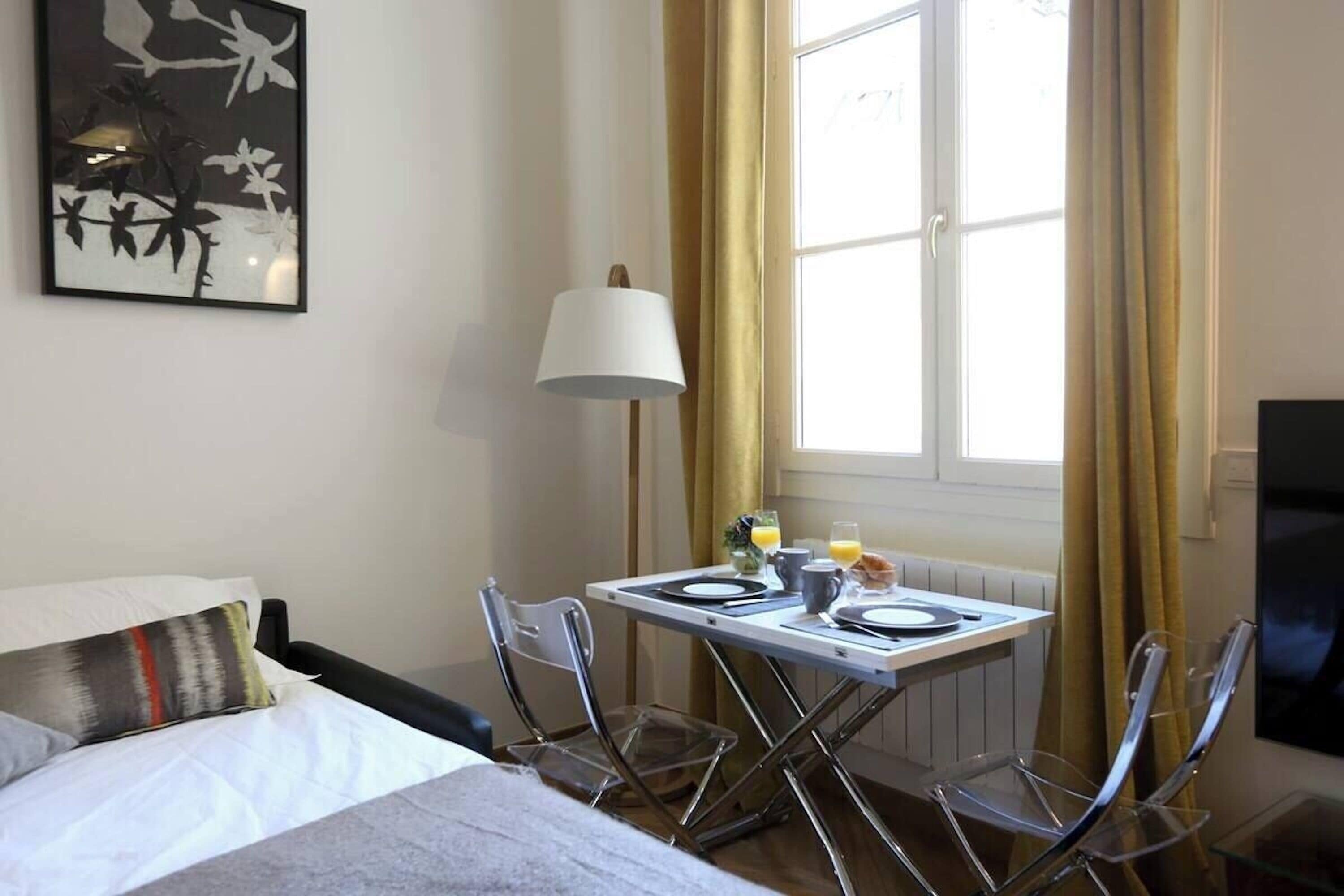 Ett hotellrum med en säng, ett matbord med stolar, en lampa och ett fönster med gardiner.