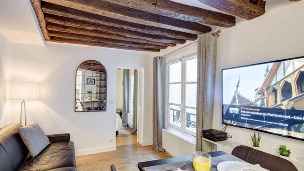 A modern living room with a wooden ceiling, a sofa, a dining table set for two, and a TV mounted on the wall displaying a news channel. - Le Marais Gem (Paris)