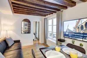 Una sala de estar moderna con techo de madera, un sofá, una mesa de comedor preparada para dos personas y un televisor en la pared mostrando un noticiero.