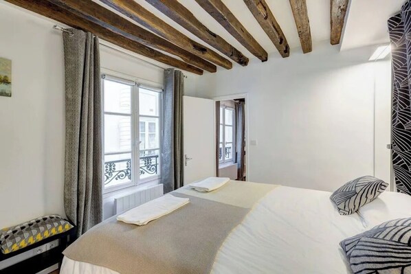 A bedroom with a large bed, wooden beams on the ceiling, a window with curtains, and a view of a balcony. - Le Marais Gem (Paris)