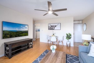 A modern living room with a flat-screen TV, a ceiling fan, a dining area with a table and chairs, and a sofa.