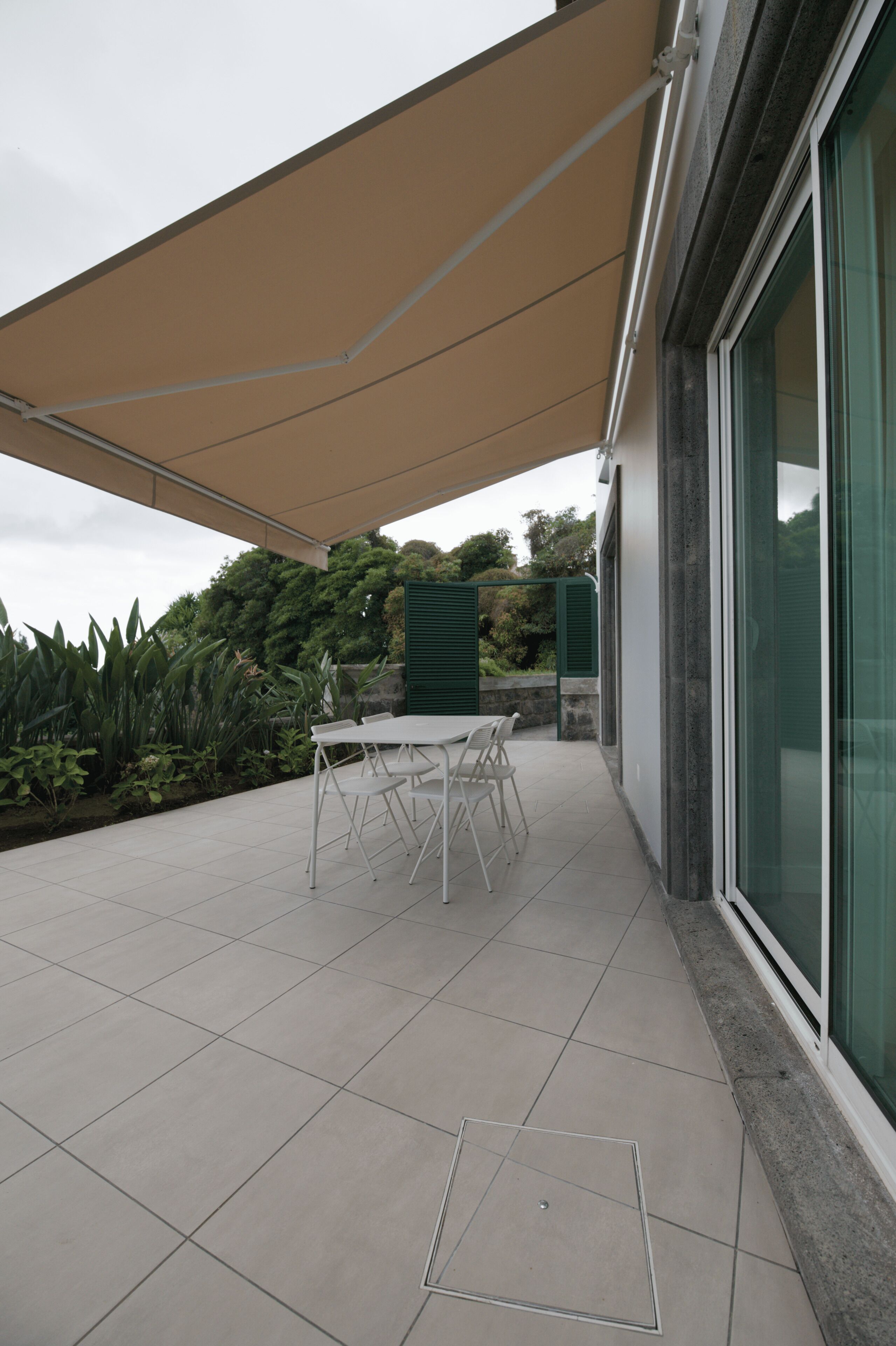 Un patio cubierto con mesa y sillas, rodeado de follaje y un edificio con amplios ventanales.