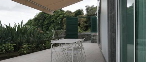 Un patio cubierto con mesa y sillas, rodeado de follaje y un edificio con amplios ventanales.