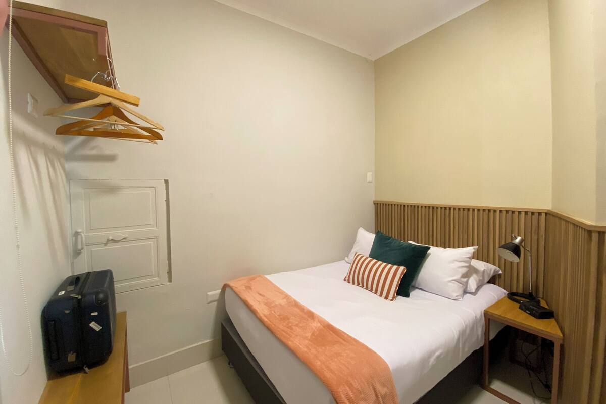 A hotel room with a bed, bedside table, lamp, and a hanging clothes rack.