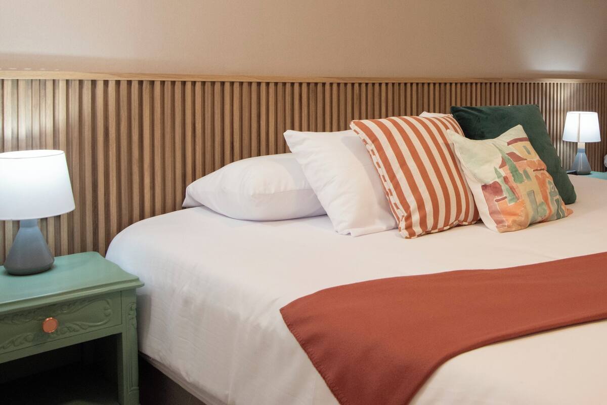 A neatly made bed with white bedding and a brown throw blanket, flanked by a green nightstand with a lamp, and a wooden headboard.