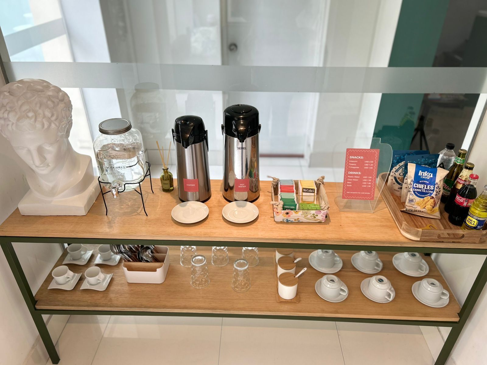 Un expositor de vidrio con una cafetera, dispensador de agua caliente y una variedad de aperitivos y bebidas.