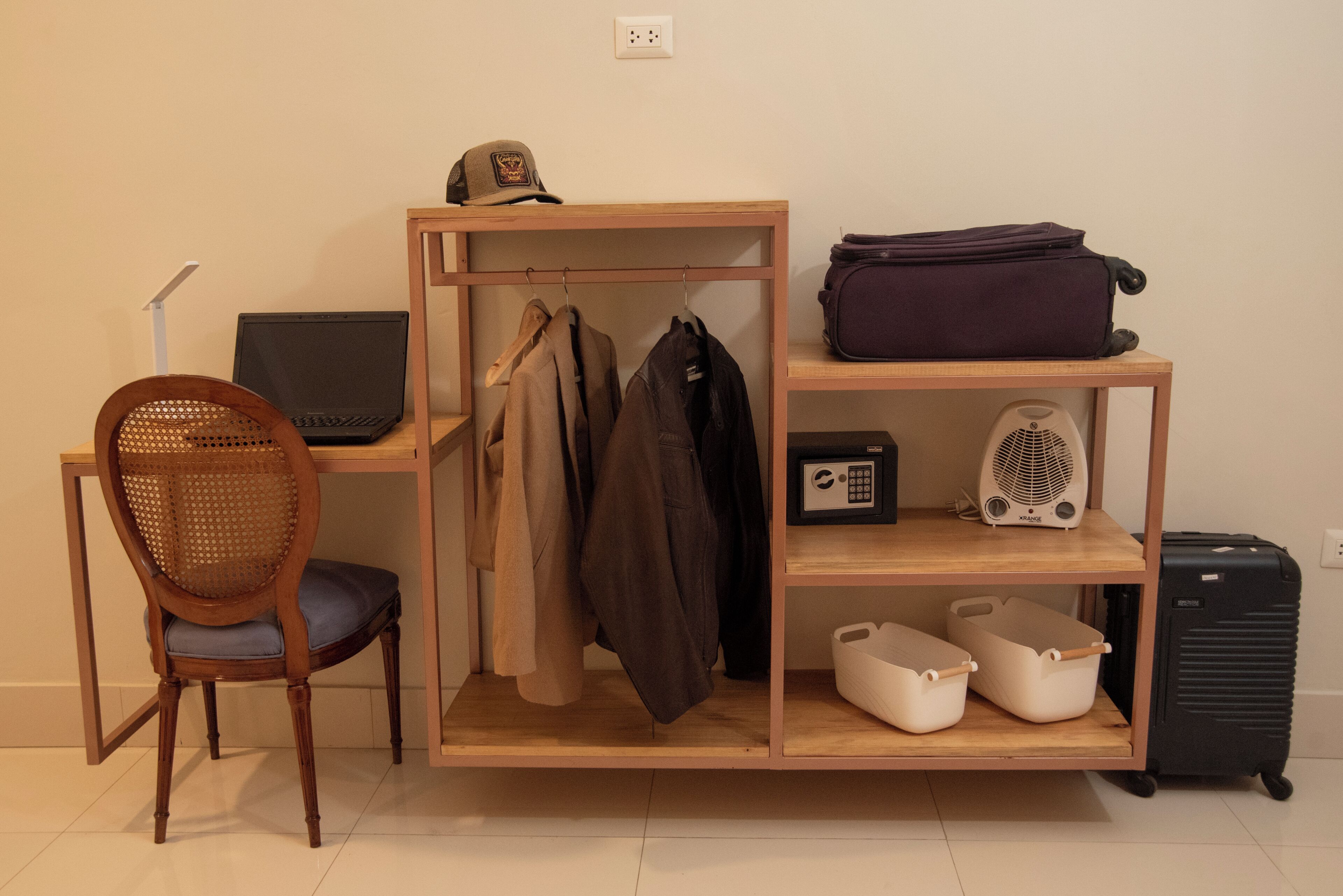 A room with a wooden desk, a laptop, a chair, a coat rack with jackets, a radio, storage bins, and a suitcase.
