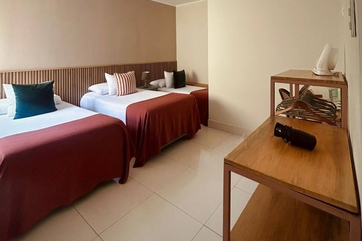 A hotel room with two beds, a wooden desk, a fan, and a shoe rack.