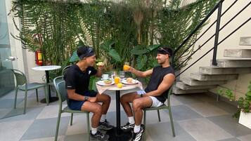 Duas pessoas sentadas em uma mesa ao ar livre com comida, cercadas por vegetação e uma escada.
