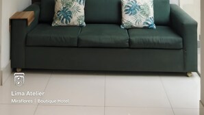 A green sofa with tropical leaf pillows, a framed abstract painting, and a small blue rabbit sculpture on a wooden side table. - Lima Atelier (Lima)
