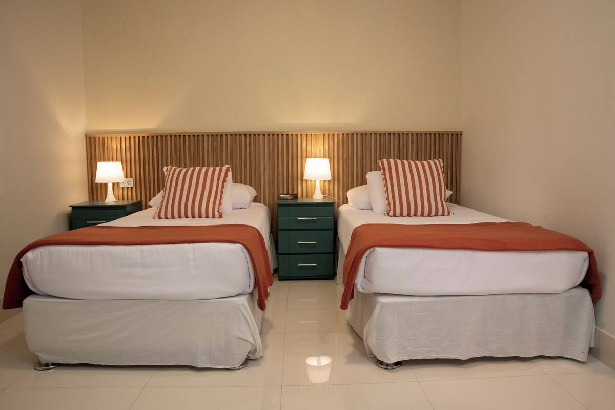 A twin room with two beds, each with a pillow and a lamp, a wooden headboard, and a green nightstand with drawers.