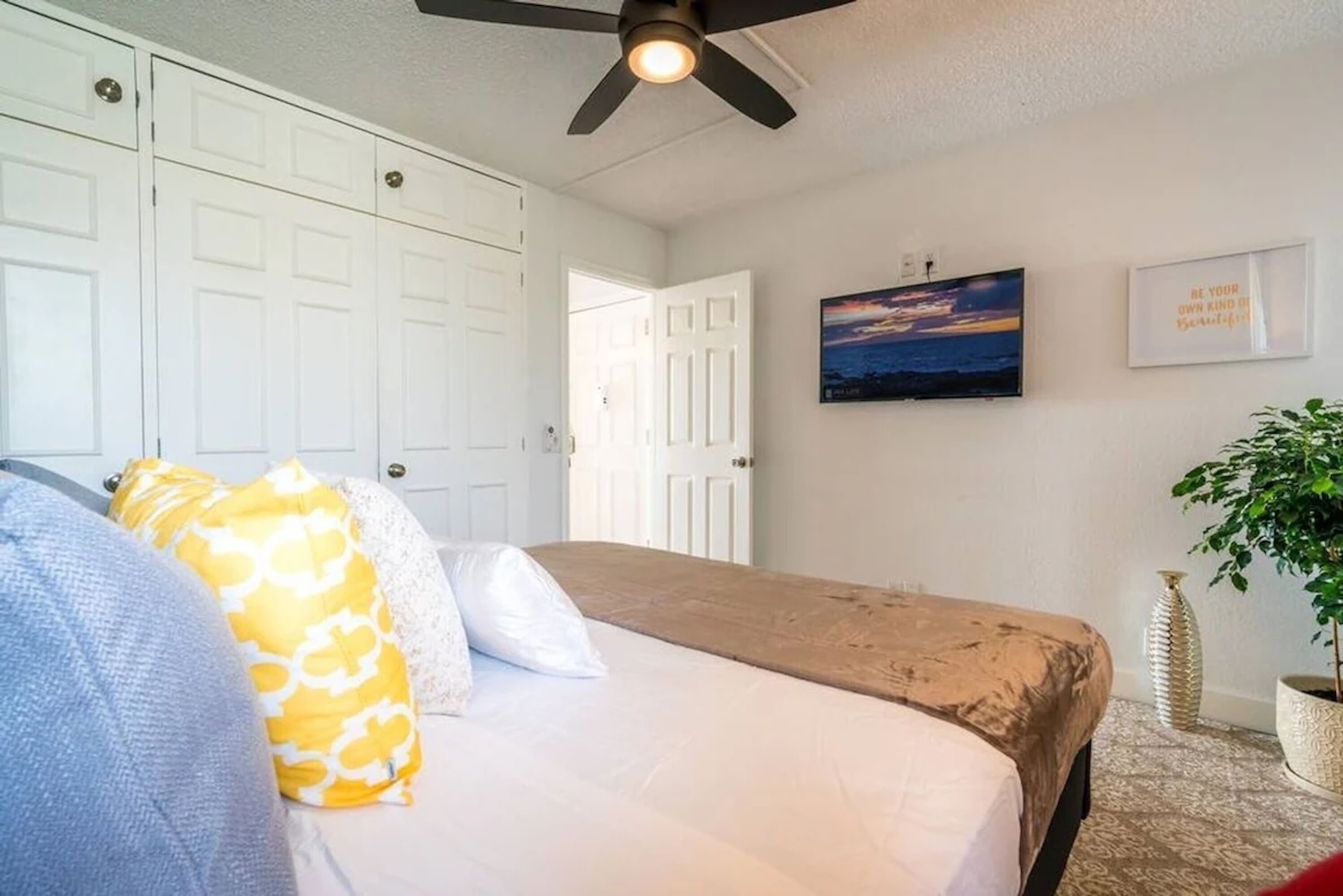 A bedroom with a bed, a ceiling fan, a wall-mounted TV, and a door leading to another room.
