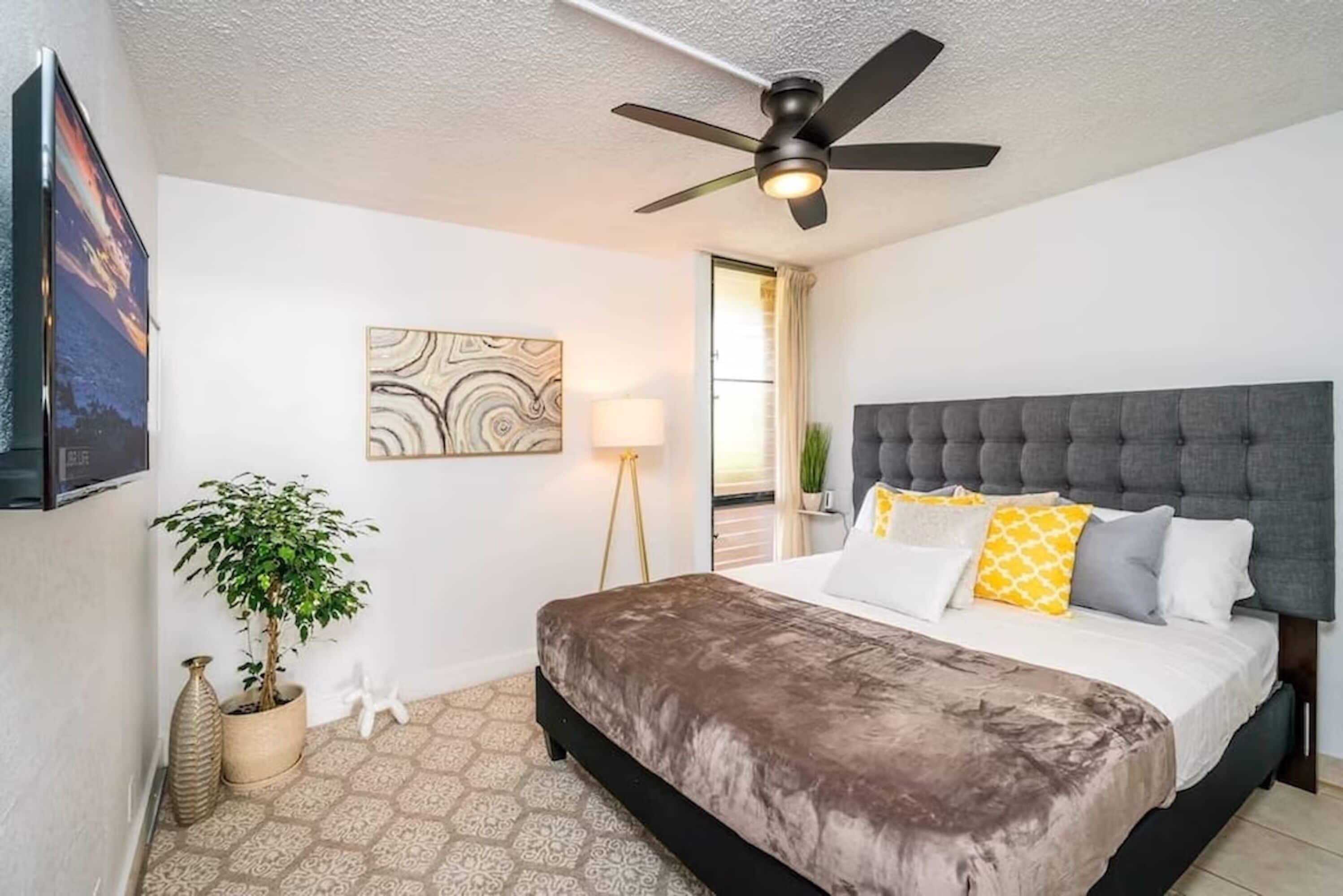 A bedroom with a bed, a ceiling fan, a wall-mounted TV, a floor lamp, a potted plant, and a decorative wall piece.