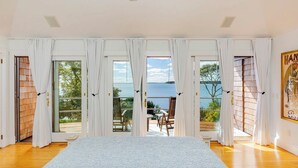 Een slaapkamer met een groot bed, witte gordijnen en uitzicht op zee door middel van glazen schuifdeuren.