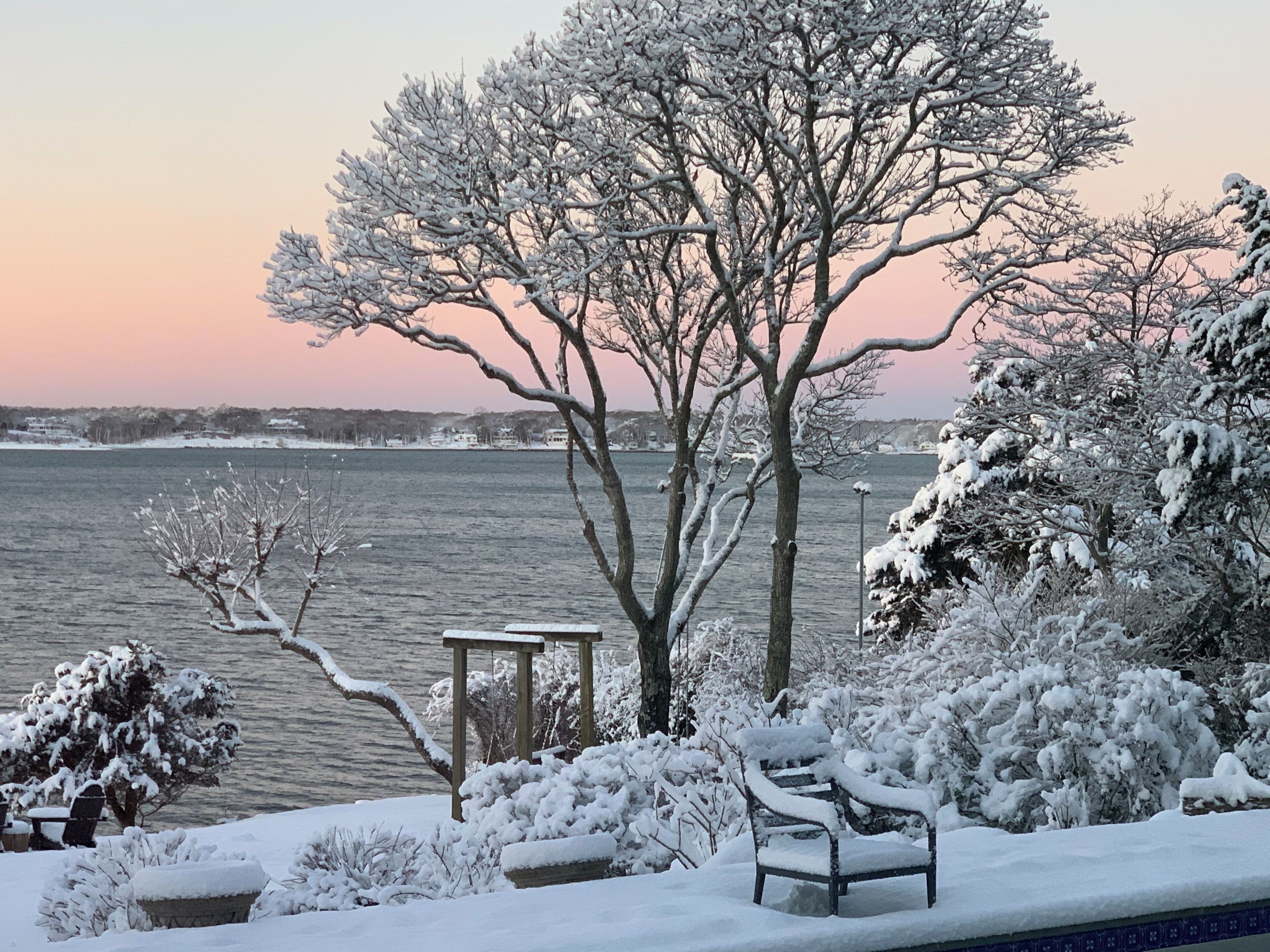 A bench and a table by a snow-covered lake with a view of the water and distant buildings.