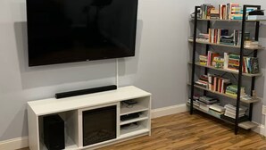 Ein Zimmer mit einem an der Wand befestigten Flachbildfernseher, einem weißen Unterstand darunter und einem schwarzen Bücherregal, das mit Büchern gefüllt ist, rechts daneben.
