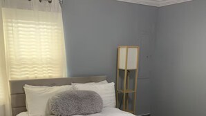 Ein ordentlich bezogenes Bett mit weißer Bettwäsche, einem grauen Zierkissen und einem Nachttisch aus Holz mit einer Lampe.